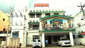 A building with a sign reading "UMA RESIDENCY" and a mural depicting a tree and figures. There are two cars parked in front, and a person is standing near the entrance. - Uma Residency (Mayiladuthurai)