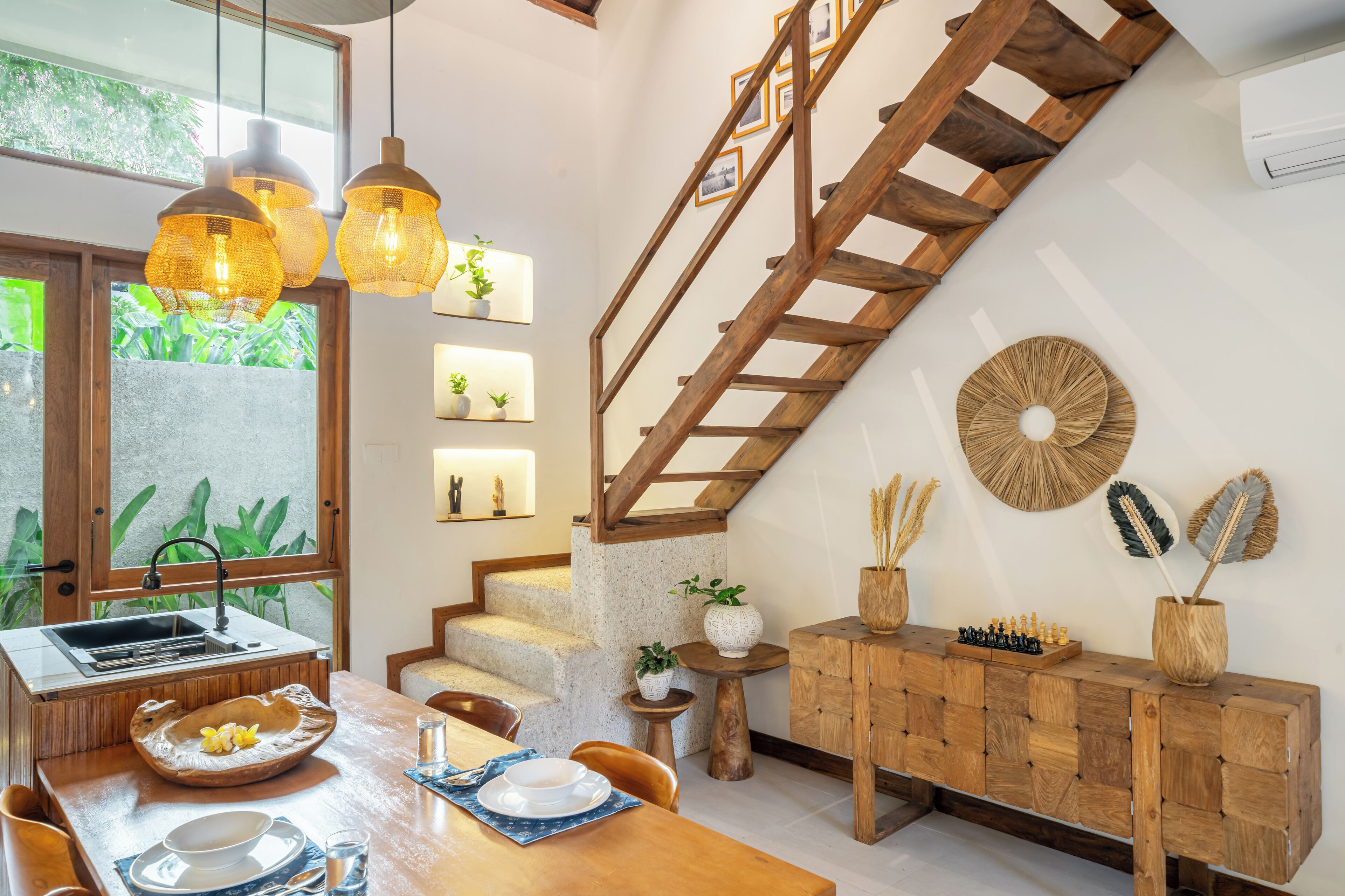 A modern kitchen with a wooden staircase, a dining area with a wooden table, and a view of greenery outside.