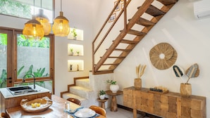 A modern kitchen with a wooden staircase, a dining area with a wooden table, and a view of greenery outside. - Chindaga Villa Canggu (Canggu)