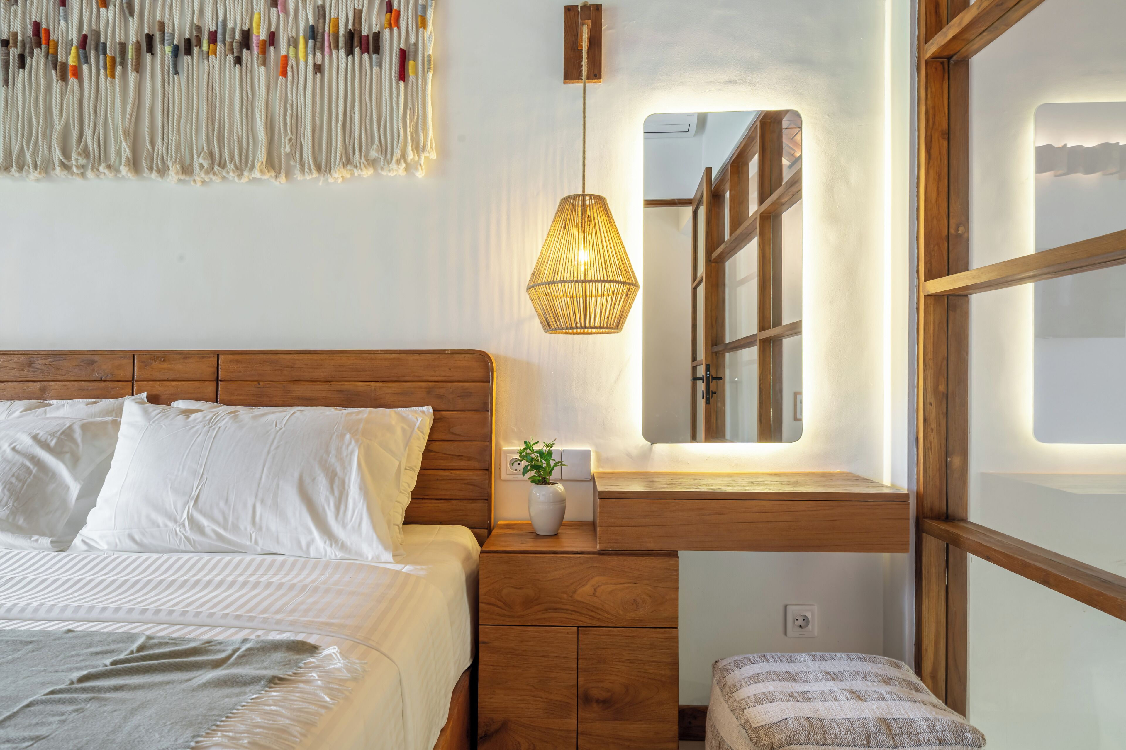 A bedroom with a wooden headboard, a bedside table with a plant, a hanging light fixture, and a large mirror reflecting the room.