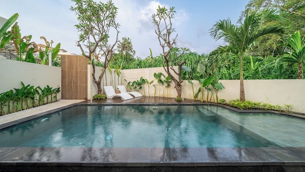 A rectangular swimming pool with a wooden deck, surrounded by tropical plants and trees. - Chindaga Villa Canggu (Canggu)