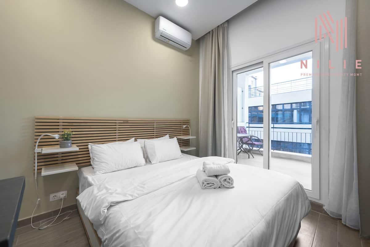 A modern hotel room with a neatly made bed, a bedside table with a lamp, and a view of the outside balcony.
