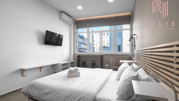 Habitación de hotel moderna con cama, televisión, ventana con vistas a edificios y un logo en la pared.