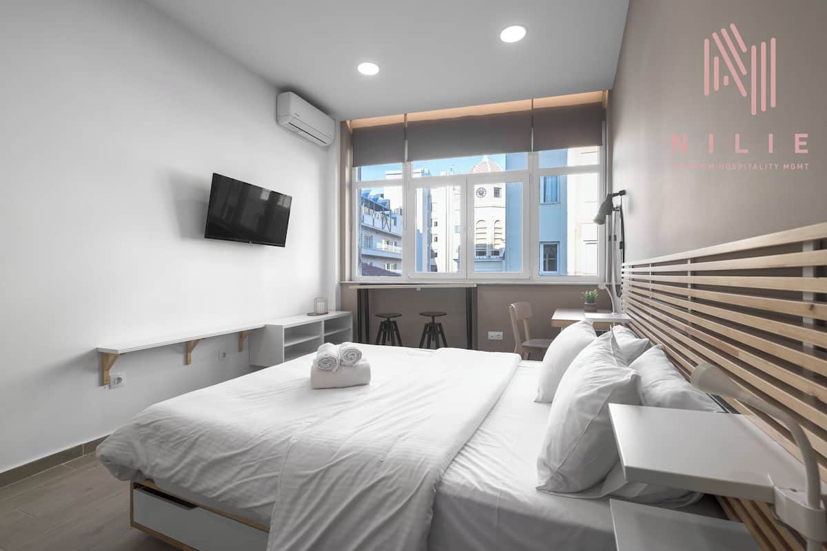 A modern hotel room with a bed, a TV, a window with a view of buildings, and a logo on the wall.