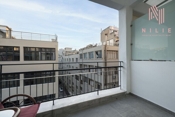 A balcony with a view of a cityscape, a glass wall with the logo 'NILIE PREMIUM HOSPITALITY MGMT', and a metal railing. - Fragkon 24 Nilie Hospitality MGMT (Thessaloniki)