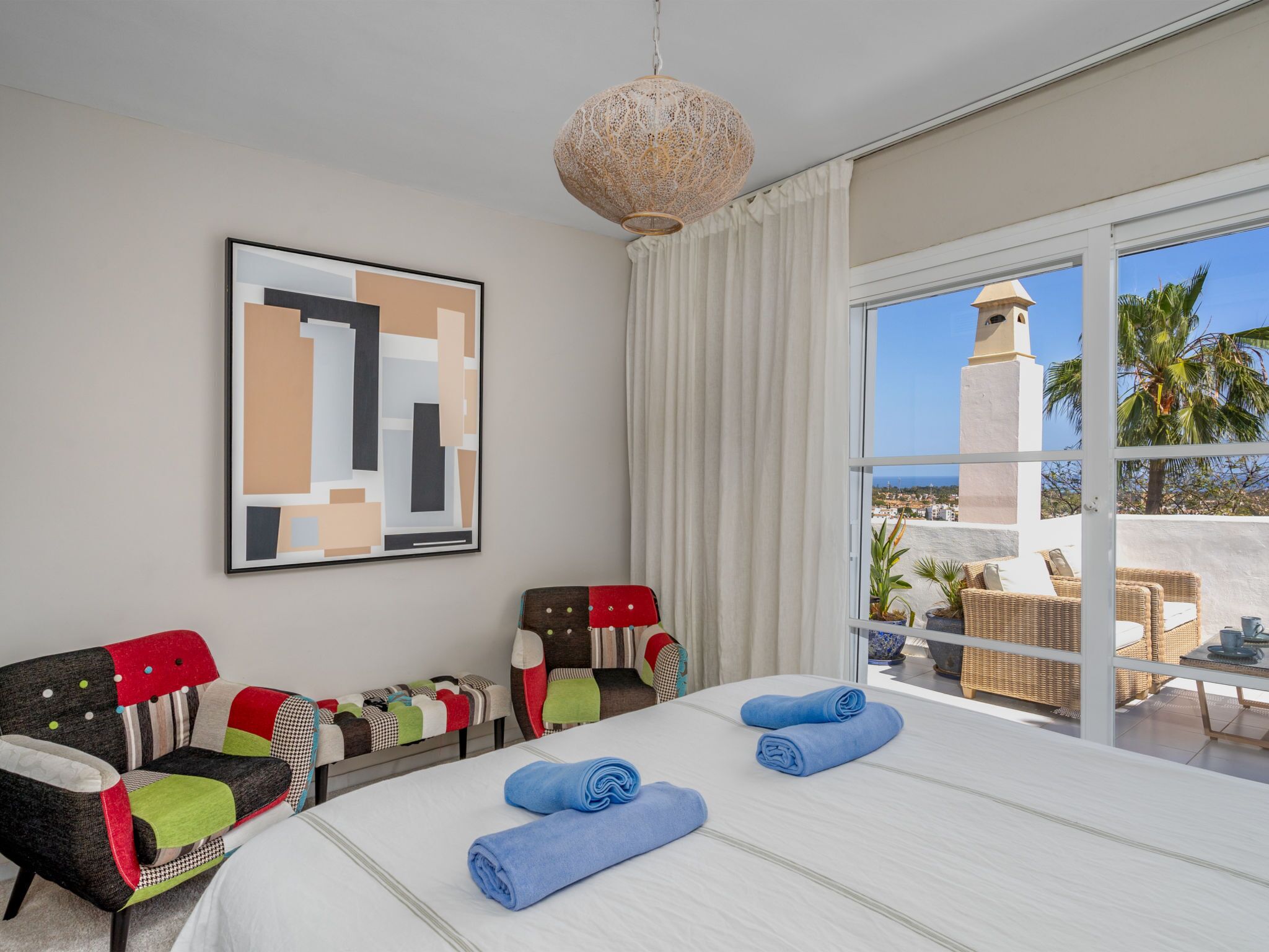 A hotel room with a bed, two chairs, a large window with a view of palm trees and a building, and a hanging light fixture.