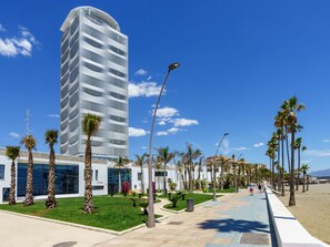 Een modern hotel met een gebogen gevel, een verzorgd gazon en een strandpromenade omzoomd met palmbomen.