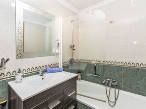Un baño con lavamanos blanco, tocador de madera oscura, un espejo grande, ducha y baldosas de mármol verde.