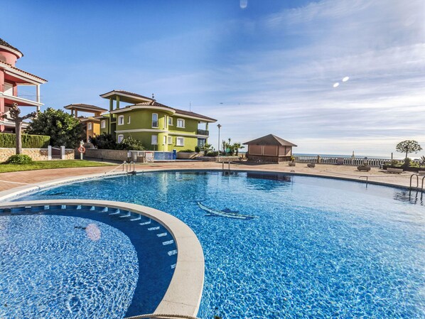 Una piscina con superficie de agua azul cristalina, rodeada por una terraza pavimentada y un muro de piedra bajo. En el fondo, edificios de varios pisos con balcones y vista al mar.