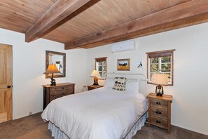 Un dormitorio con techo de madera, una cama con ropa de cama blanca, dos mesitas de noche con lámparas y una ventana con vista a árboles.