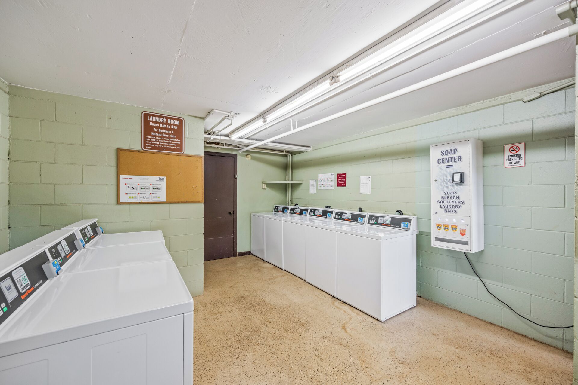 Ein Waschraum mit mehreren Waschmaschinen, einer Seifenstation und Schildern, die Öffnungszeiten und Rauchverbot angeben.