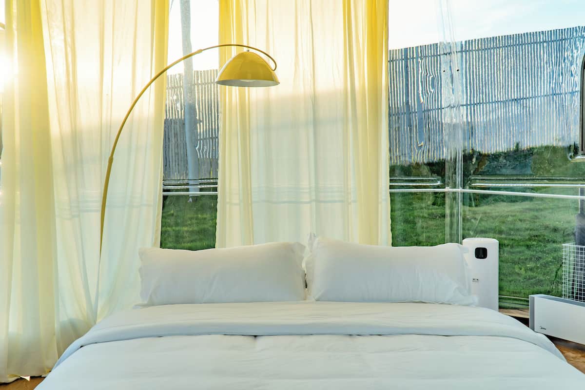 A bedroom with a bed, white curtains, a floor lamp, and a view of greenery outside.