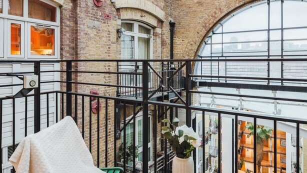 A high-ceilinged room with exposed brick walls, large arched windows, and a wooden table with a white cloth and a vase of flowers.