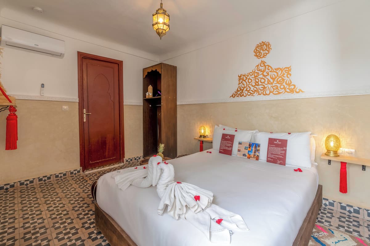A hotel room with a large bed, a wooden wardrobe, a wall-mounted air conditioner, a decorative wall piece, and a small shelf with a lamp.