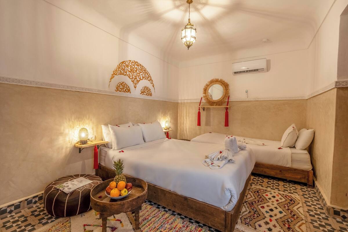 A hotel room with two beds, a fruit bowl, a small table with a lamp, and decorative wall art.