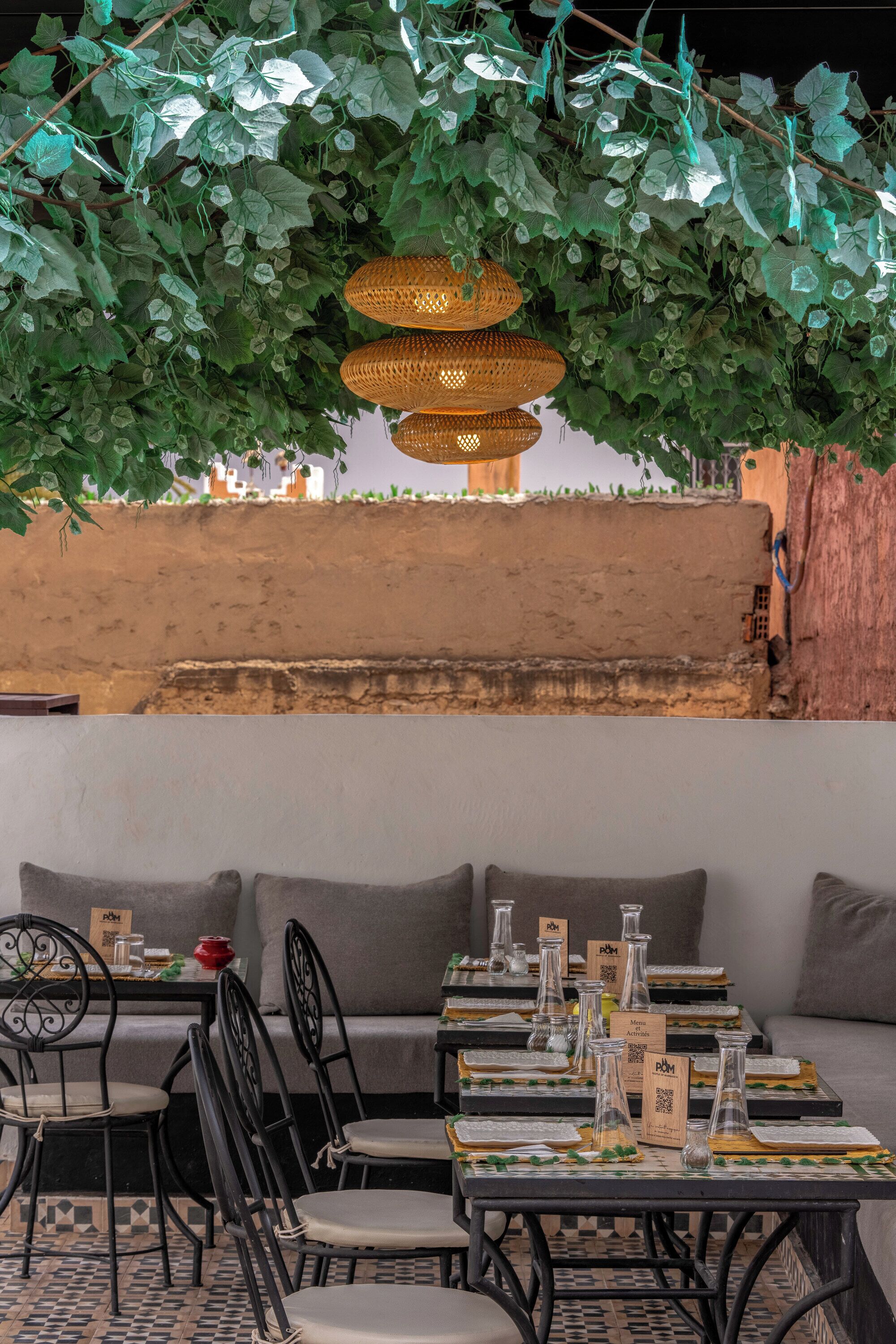 Un área de comedor al aire libre con mesas dispuestas para dos personas, rodeada de asientos acolchados y follaje verde colgante.