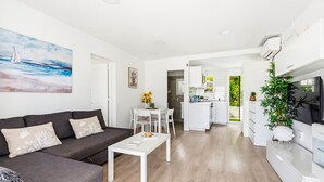 A modern living room with a sofa, coffee table, and artwork. A kitchen is visible in the background with white cabinets and a dining area. - Castellsol 315 (Arenal d´en Castell)