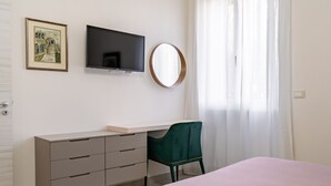 Une chambre à coucher comprenant un lit, un bureau avec des tiroirs, une chaise, un téléviseur, un tableau encadré et une fenêtre avec des rideaux.