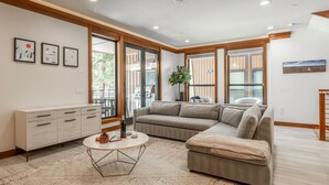 A modern living room with a sectional sofa, a round coffee table, and a cabinet. There are large windows with wooden frames, and artwork on the walls. - Private Hot Tub | Near Eagle Lodge (Mammoth Lakes)