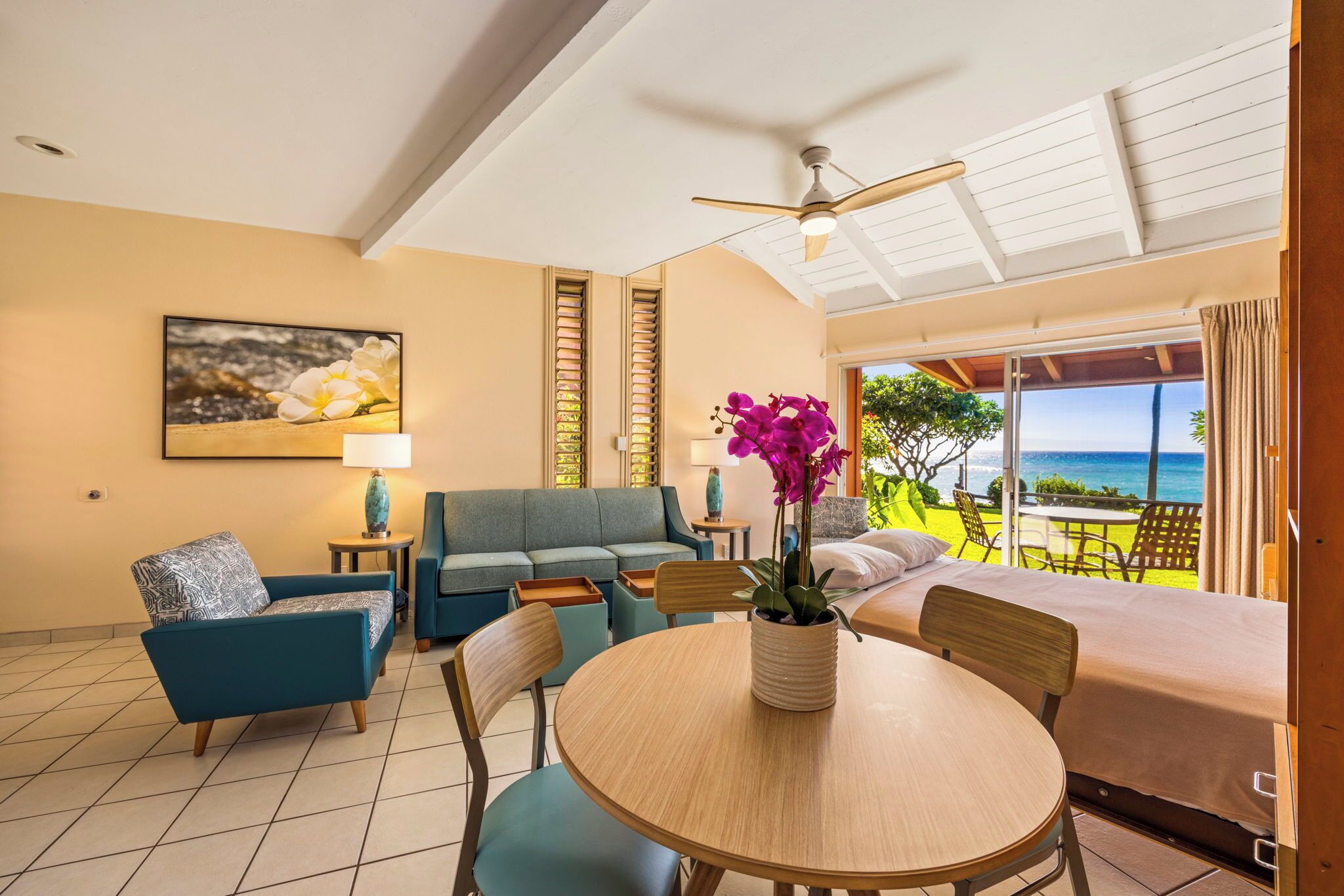 A room with a sofa, a dining table, and a bed. There is a view of the ocean through large windows.