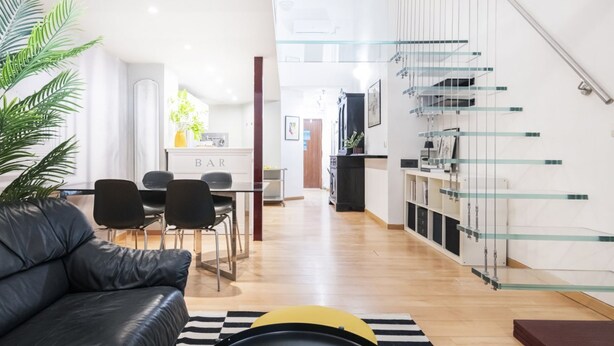 A modern living room with a black leather sofa, a dining area with a white table and chairs, and a kitchen with a glass staircase. - Easylife - Modern Comfort in Porta Venezia (Milano)