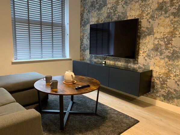 A modern living room with a round wooden table, a sofa, a television mounted on a textured wall, and a window with blinds.