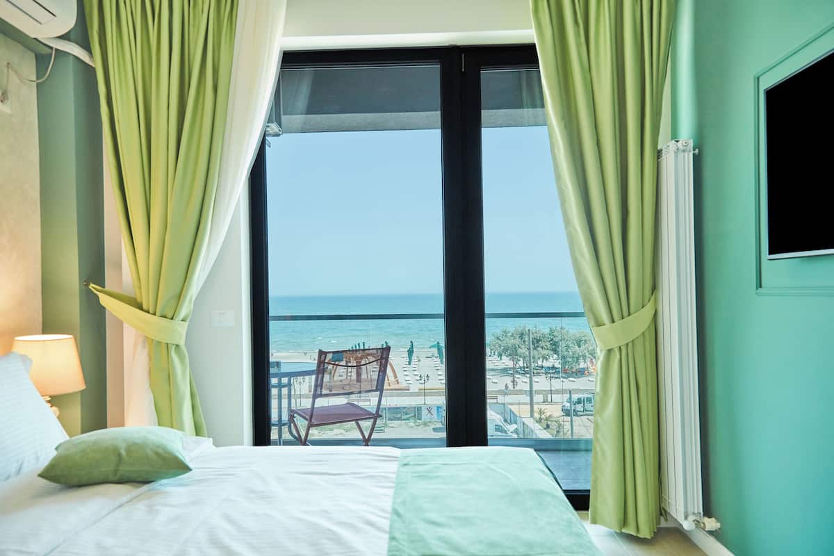 A hotel room with a bed, a chair, a view of the beach, and a window with curtains.