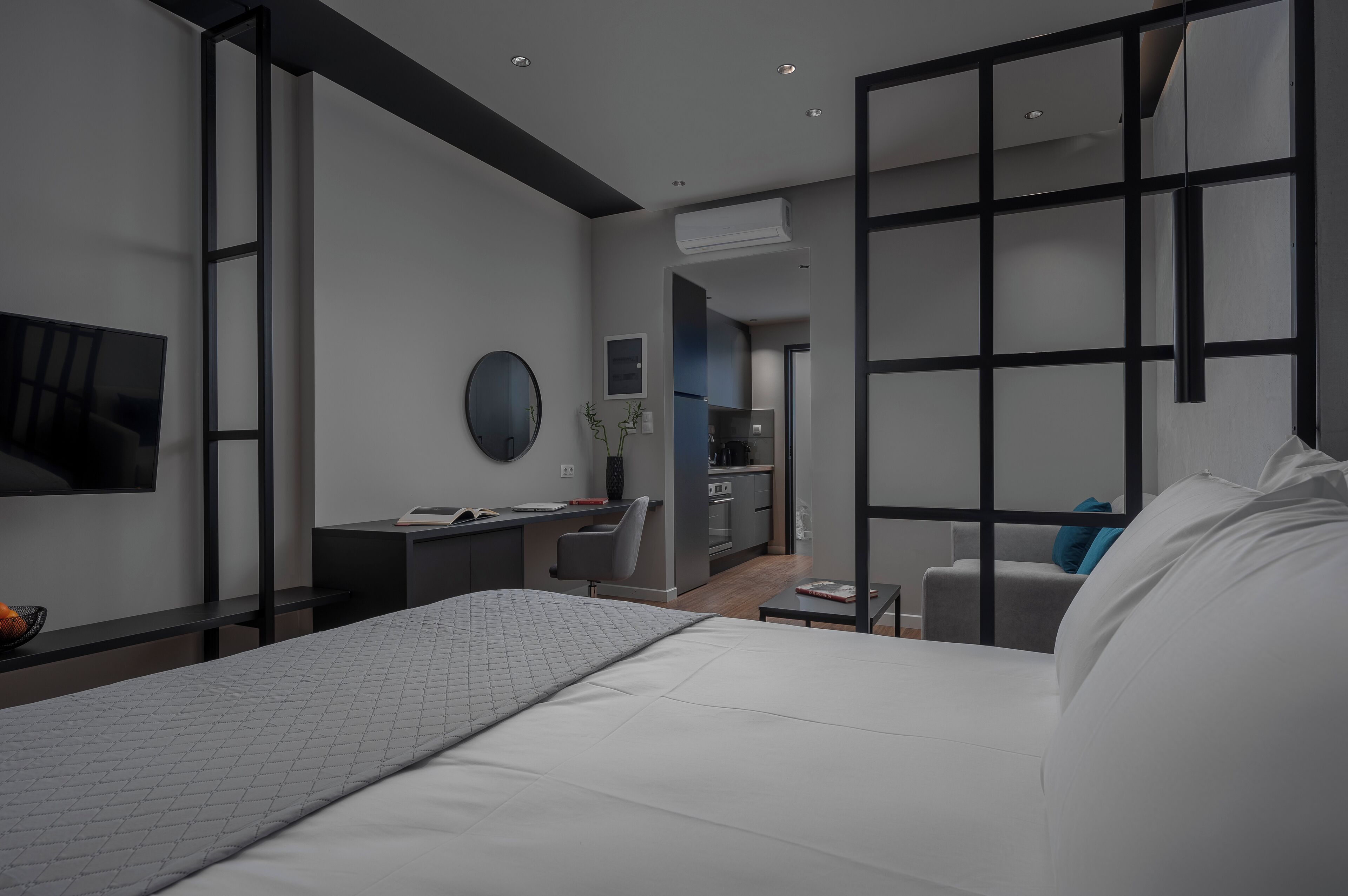 A modern hotel room with a large bed, a desk, a chair, a TV, and a kitchen area.