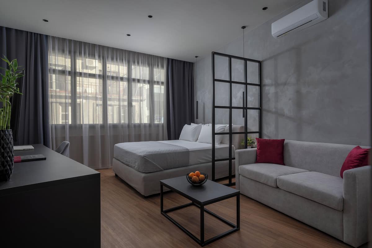 Ein modernes Hotelzimmer mit großem Fenster, einer Couch, einem Bett, einem Schreibtisch und einem kleinen Tisch mit Obstschale.