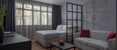 Una habitación de hotel moderna con un ventanal grande, un sofá, una cama, un escritorio y una pequeña mesa con un frutero.