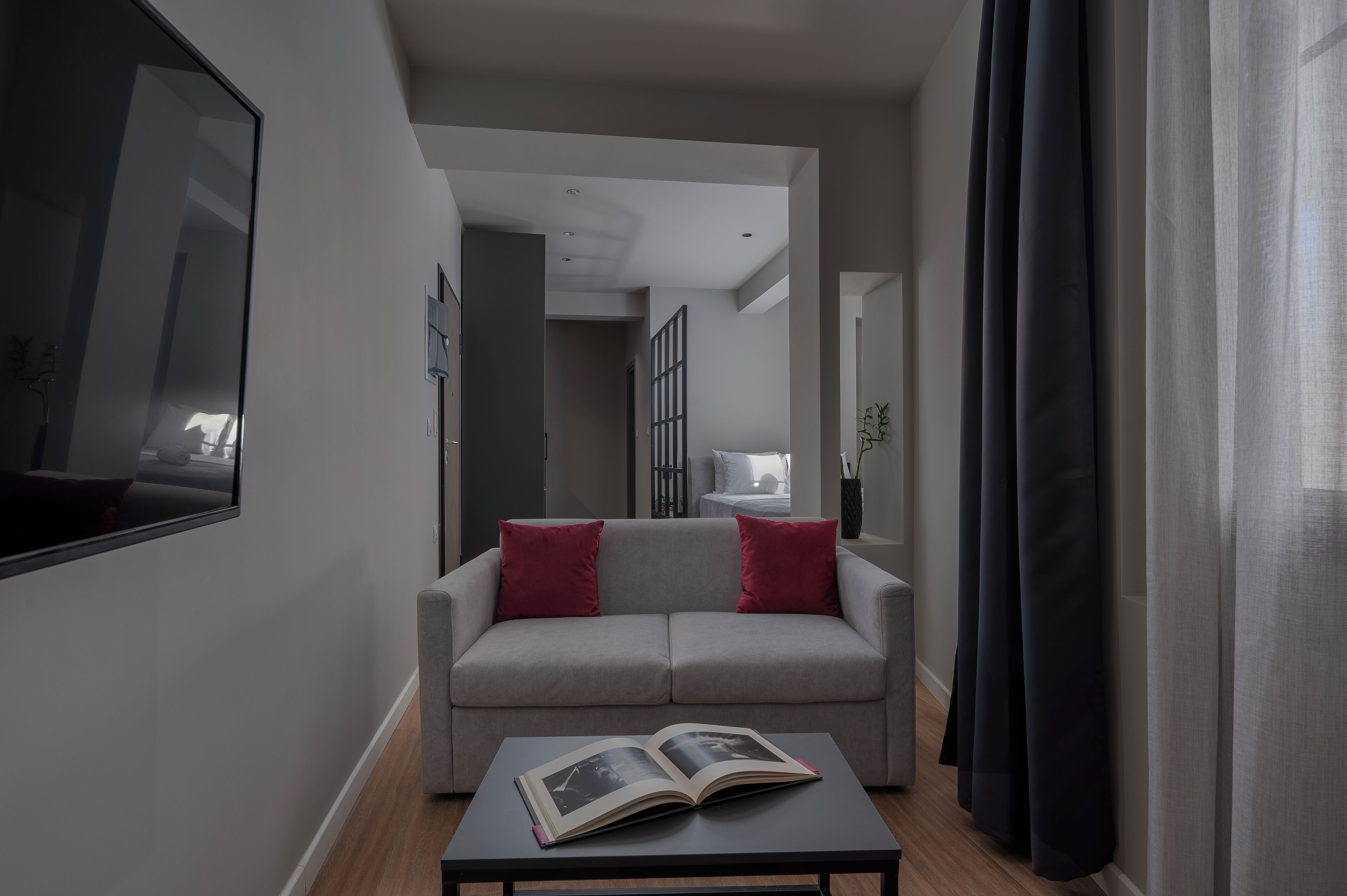 A modern living room with a grey sofa, red cushions, a coffee table with an open book, and a large window with dark curtains.