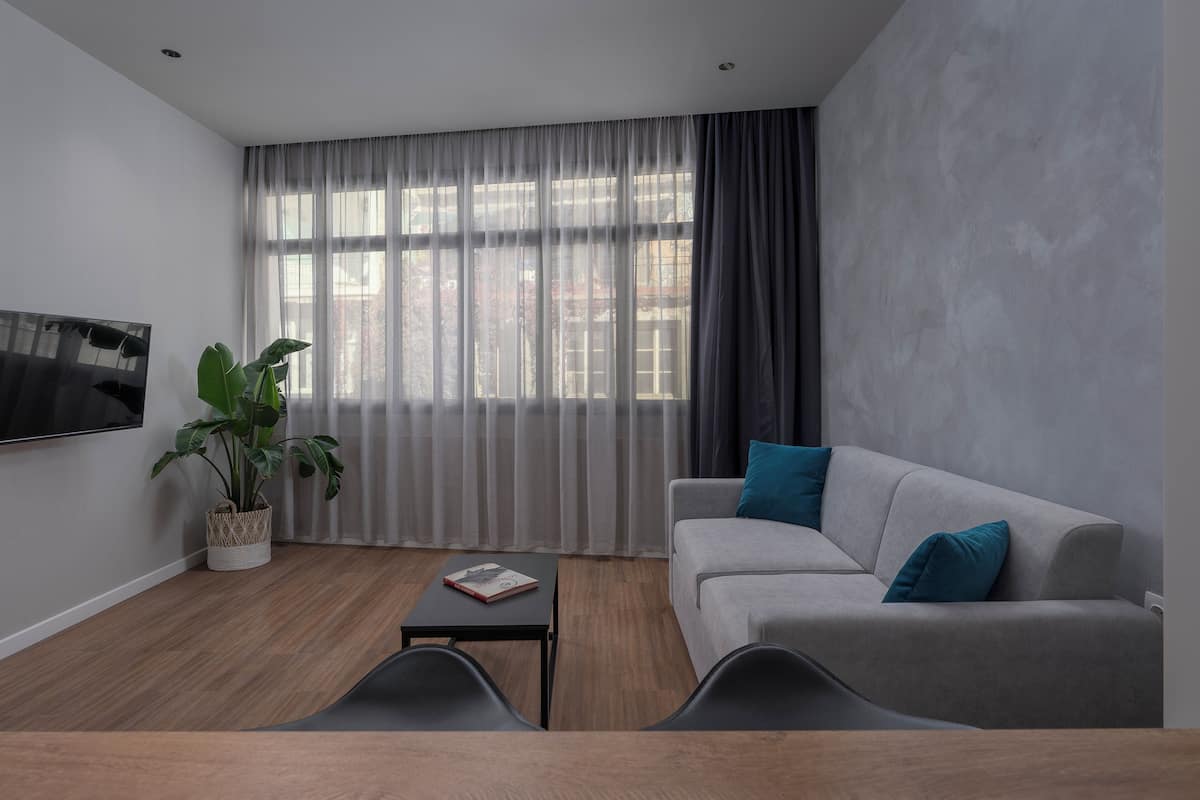 Ein modernes Wohnzimmer mit einem grauen Sofa, einem Couchtisch, einem an der Wand montierten Fernseher und einem großen Fenster mit transparenten Vorhängen.