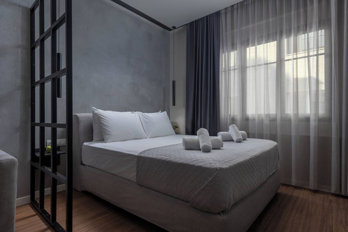 Ein modernes Schlafzimmer mit einem großen Bett, einem Nachttisch und einem Fenster mit transparenten Vorhängen.