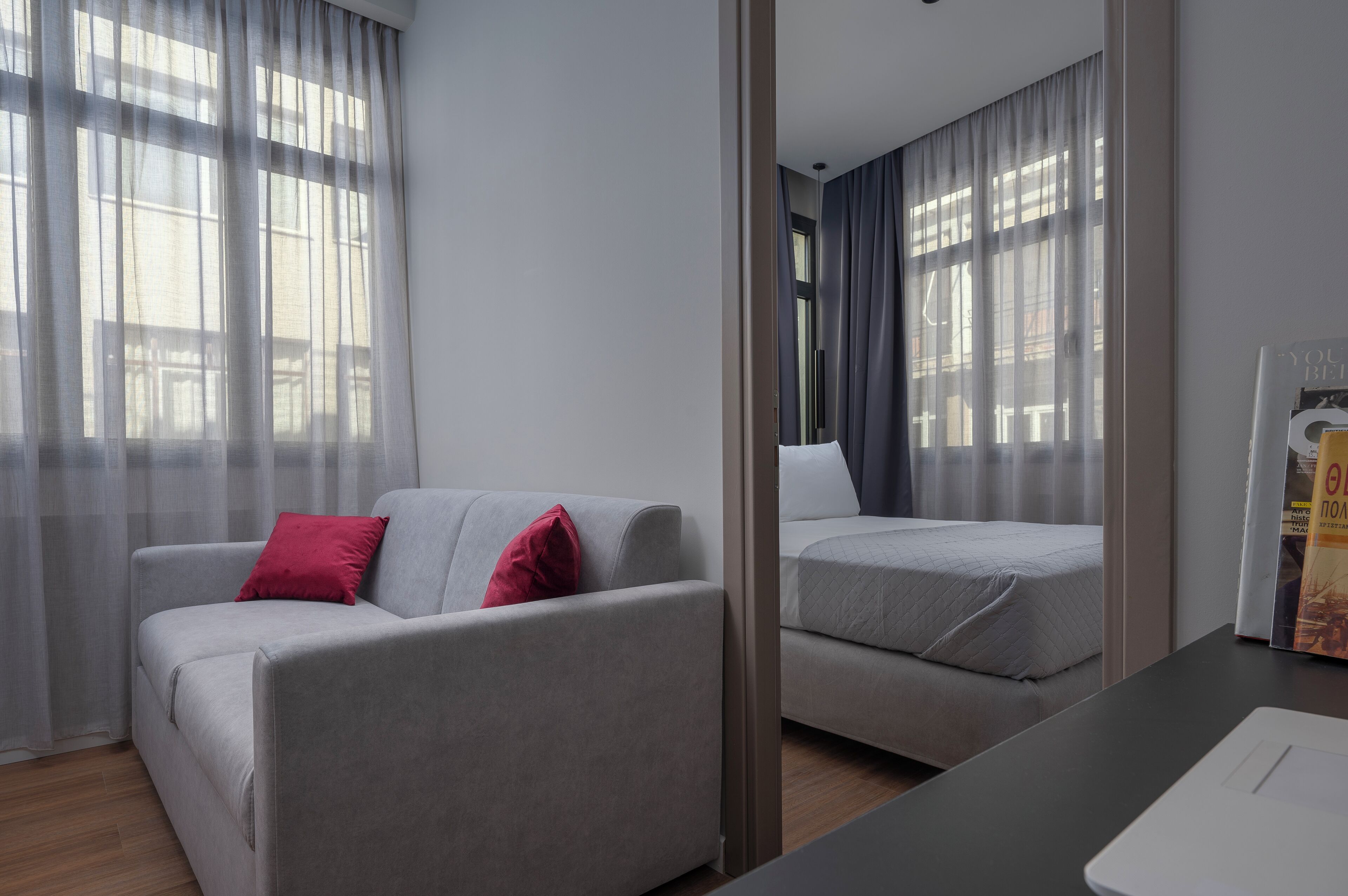 Habitació d'hotel moderna amb un sofà gris, un llit i una finestra gran amb cortines lleugeres.