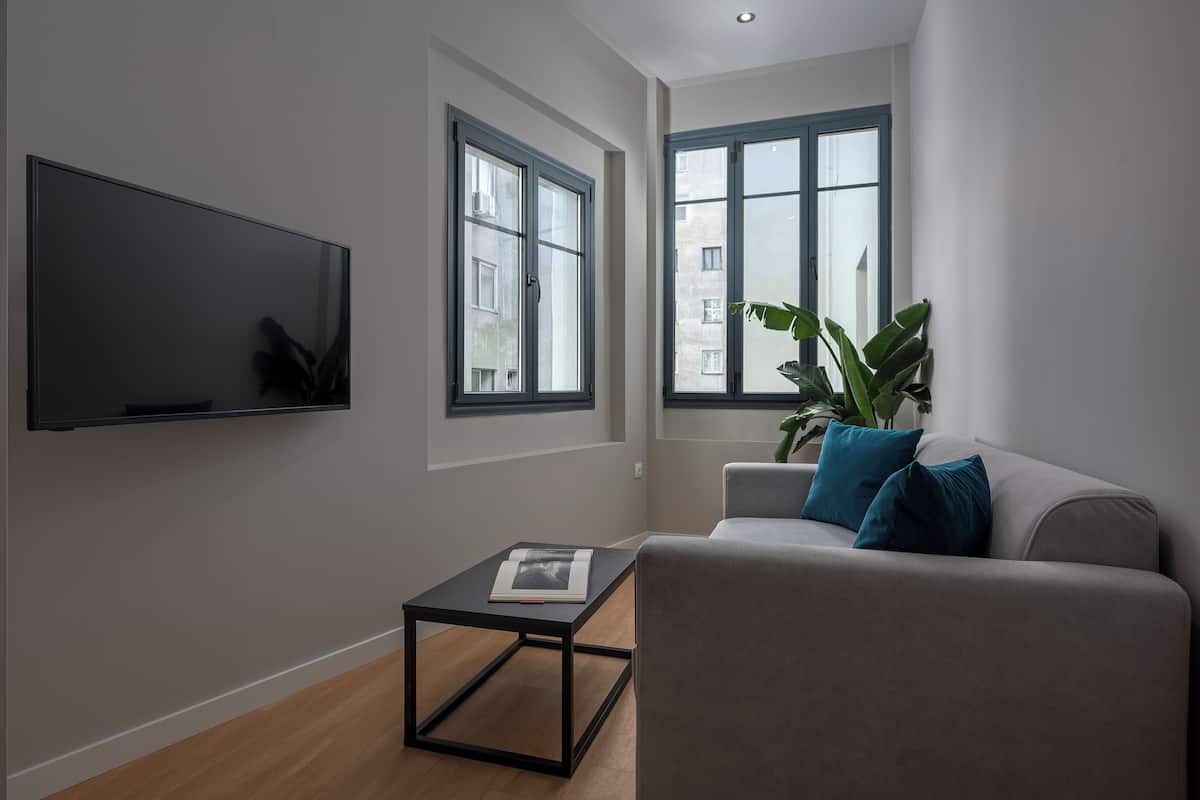 Ein modernes Wohnzimmer mit Flachbildfernseher, einem grauen Sofa mit blauen Kissen, einem kleinen schwarzen Couchtisch und großen Fenstern mit blauen Rahmen.