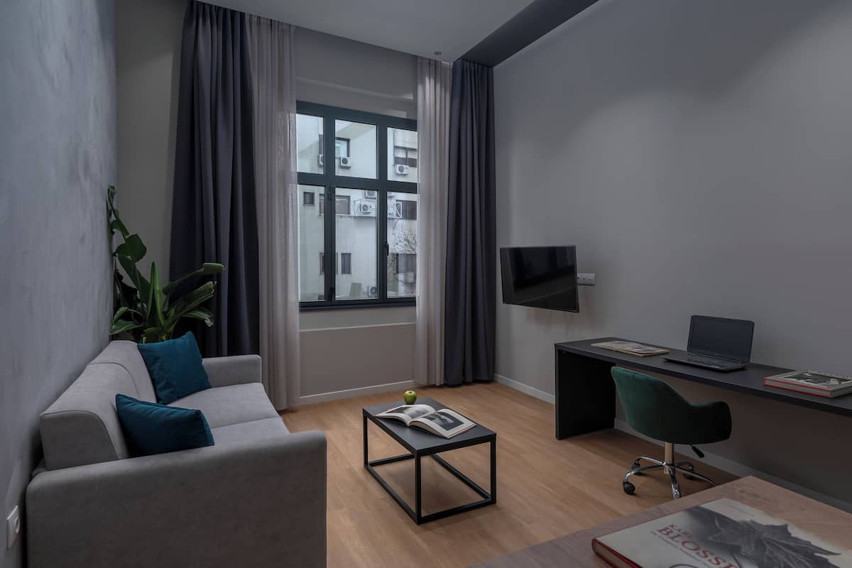Ein modernes Wohnzimmer mit einem grauen Sofa, einem kleinen Couchtisch mit einem Buch und einem Apfel, einem Schreibtisch mit einem Laptop und einem Buch sowie einem großen Fenster mit Vorhängen.