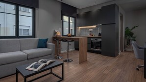 A modern living room with a grey sofa, a coffee table with a book, and a kitchen area with a fridge, oven, and bar stools. - Hebrard Industrial Suites (Thessaloniki)