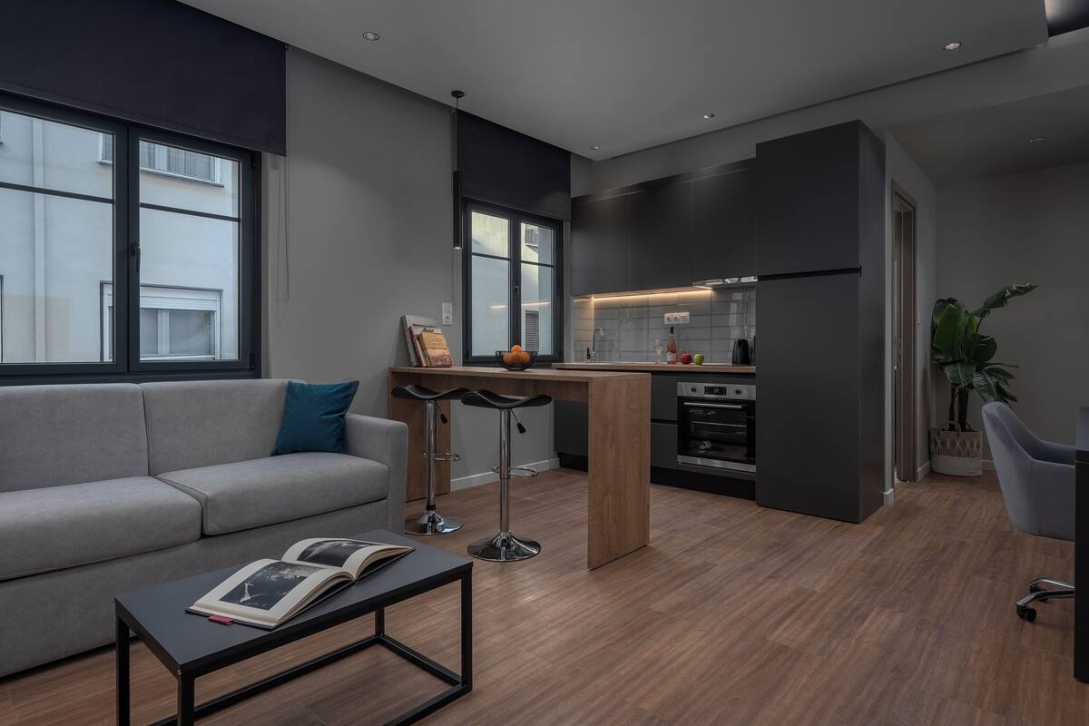 Ein modernes Wohnzimmer mit einem grauen Sofa, einem Couchtisch mit einem Buch und einer Küchenzeile mit Kühlschrank, Backofen und Barhocker.