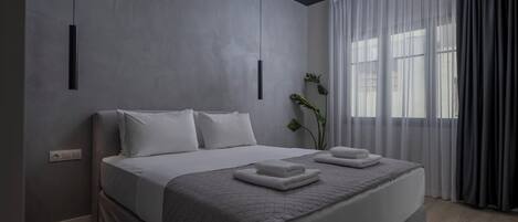 A modern bedroom with a large bed, a bedside plant, and a view of the outdoors through sheer curtains.
