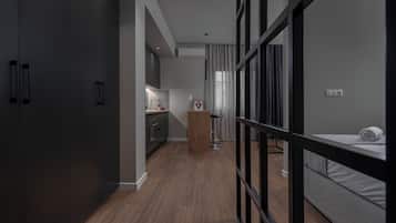A modern hotel room with a wooden floor, a bed, a desk with a chair, and a kitchenette in the background.