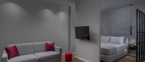A modern living room with a grey sofa, a red ottoman, a coffee table, and a flat-screen TV mounted on the wall.