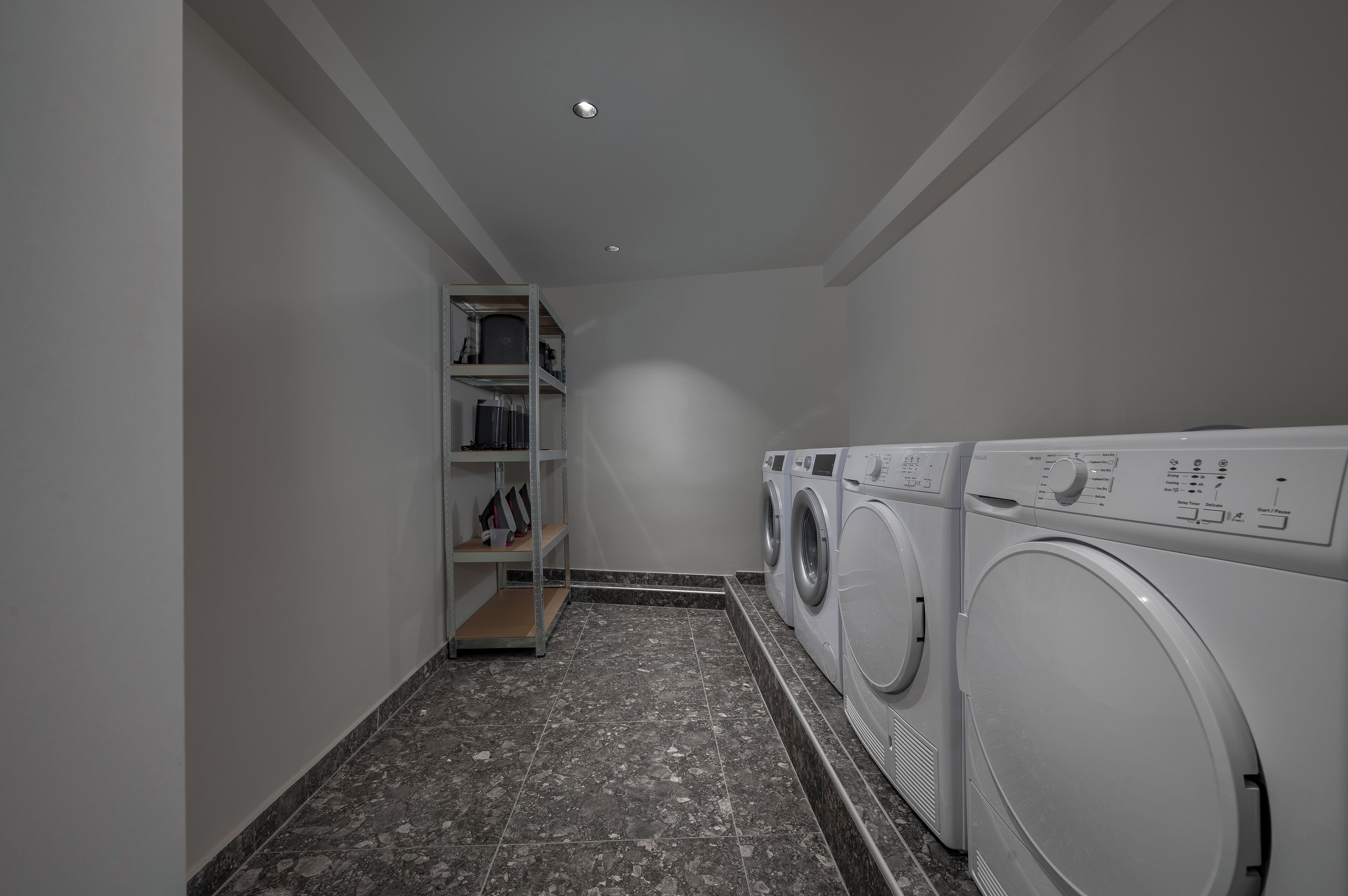 Een wasruimte met een wasmachine en droger, een plank met opgevouwen kleding en een betegelde vloer.