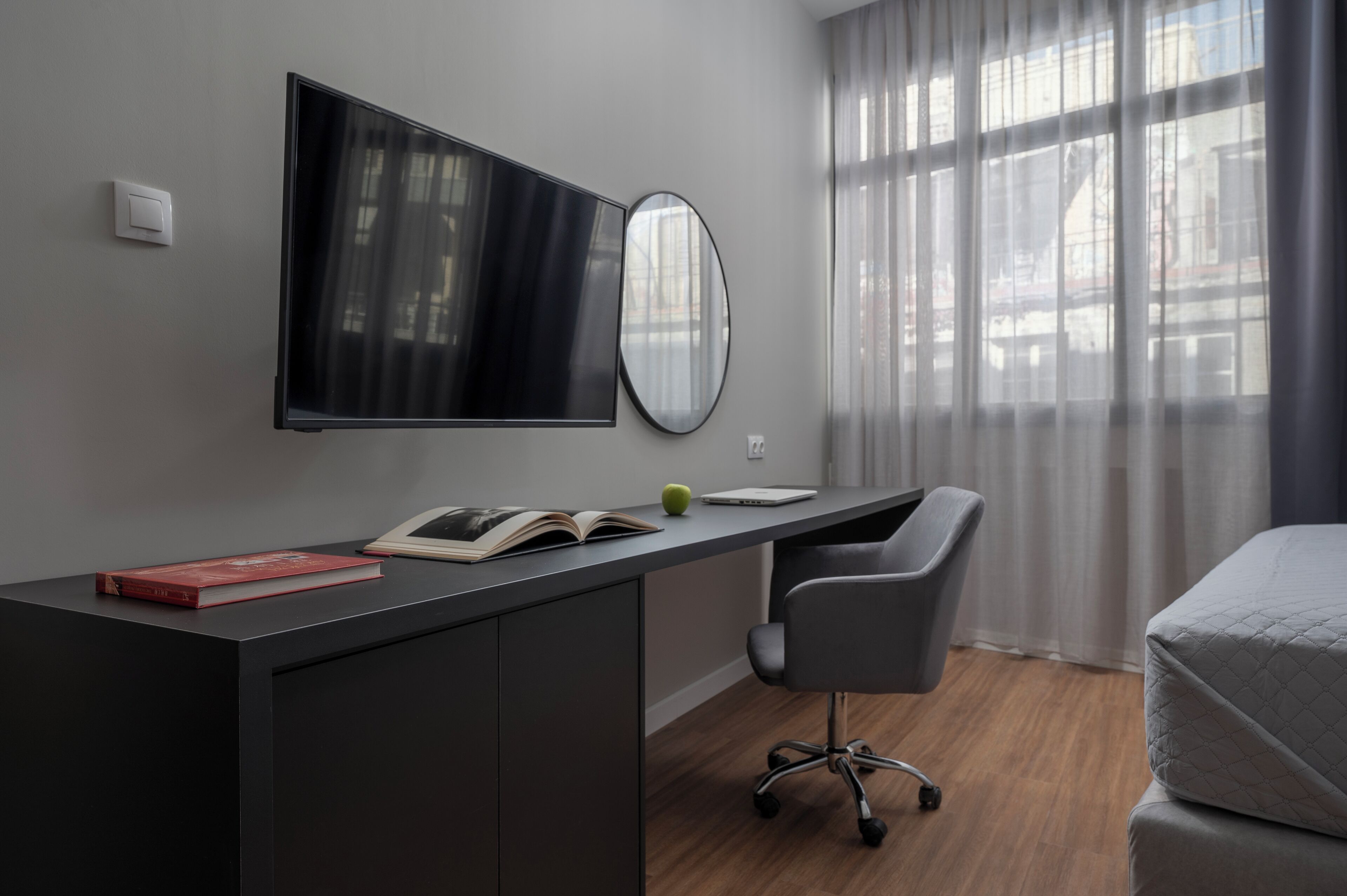 A modern hotel room with a large flat-screen TV mounted on the wall, a desk with a laptop, a chair, and a bed with a quilted cover.