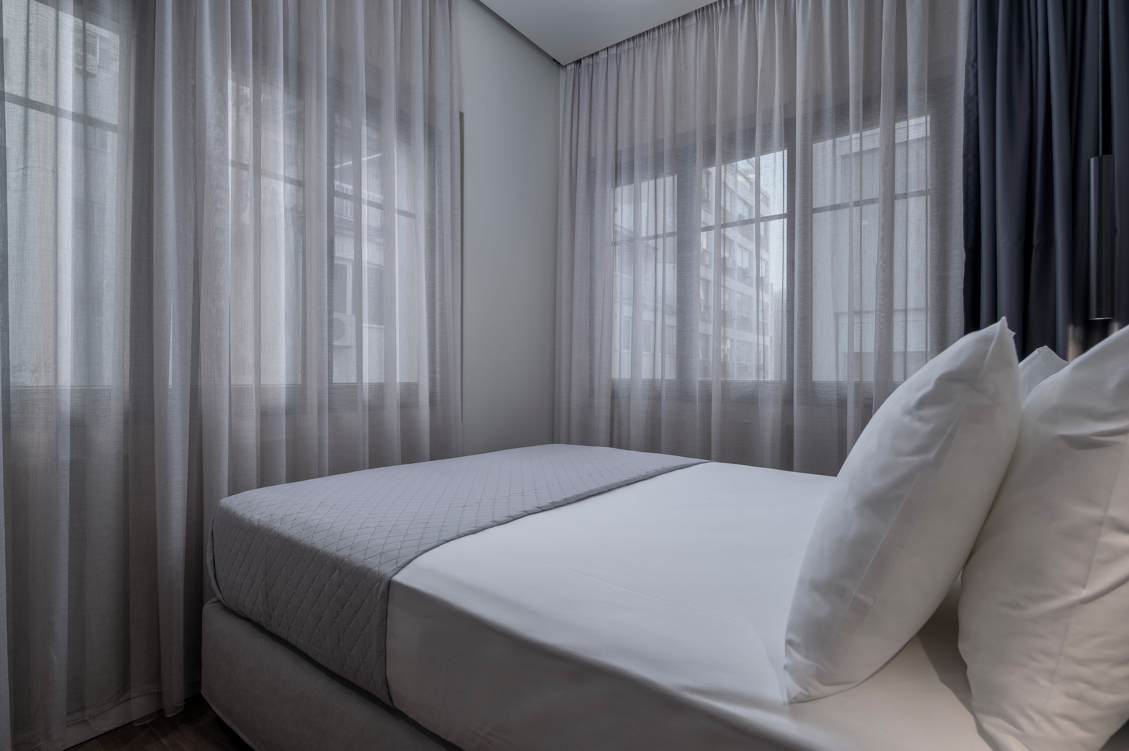 Habitación de hotel con cama, cortinas translúcidas y vistas a edificios a través de las ventanas.