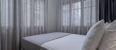Habitación de hotel con cama, cortinas translúcidas y vistas a edificios a través de las ventanas.