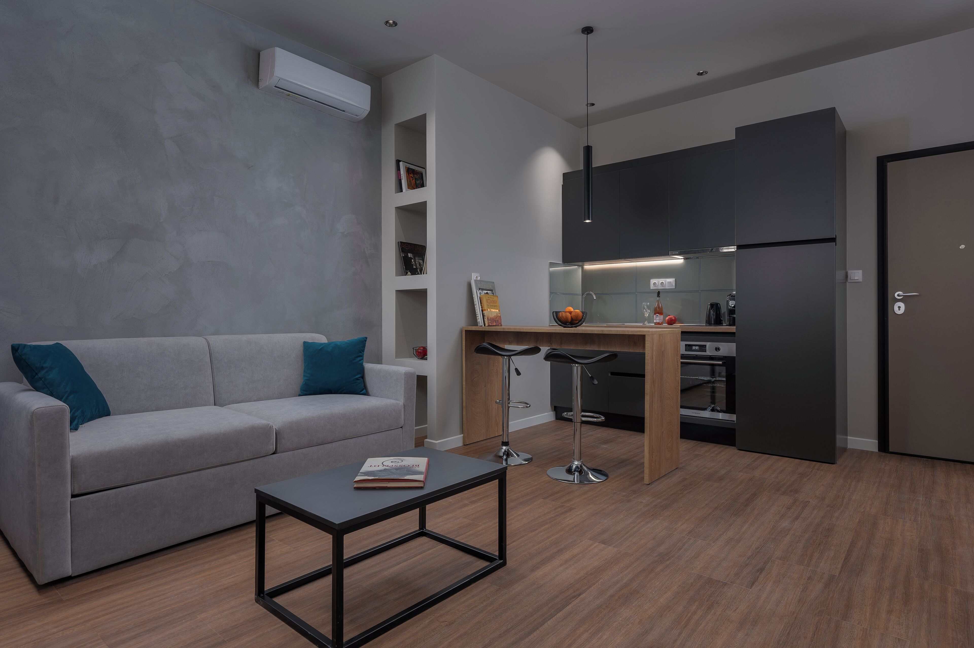 A modern living room with a grey sofa, a coffee table, and a kitchen area with a fridge, oven, and bar stools.