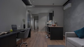 A modern, minimalist living space with a grey sofa, a desk with a laptop, and a kitchen area in the background. - Hebrard Industrial Suites (Thessaloniki)