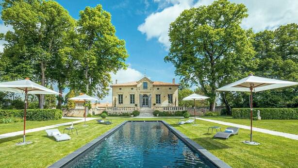 A pool with a stone border, a building with a tiled roof, and trees in the background.