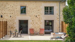 A stone building with a patio area featuring two tables, two chairs, a lounge chair, and a hot tub.