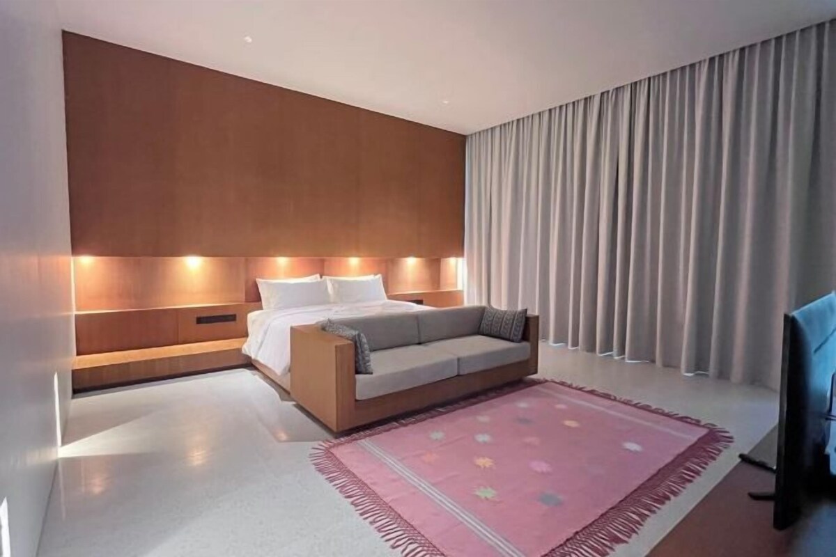 Quarto de hotel moderno com cama, sofá, televisão e uma janela grande com cortinas.